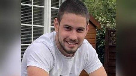 A photo of Brandon Fielder smiling towards the camera in a white t-shirt sat in a garden with a shed and some windows behind him.