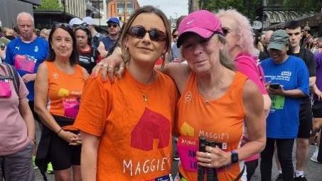 Angela (right) stands with her daughter Josie in the middle of a crowd. Josie wears and orange T-shirt with Maggie's written on the front in white letters. She wears sunglasses and her dark blonde hair is pulled back loosely from her face.