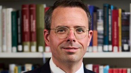 Dr Toby Simpson is standing in front of shelves of books. He is wearing a dark suit and glasses.