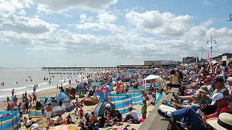 A packed seafront during a sunny day. Hundreds of people sit on the beach with umbrellas and wind shields with some swimming in the sea, and others sitting behind a sea wall on deck chairs .