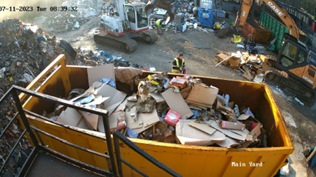CCTV of the waste yard. A large skip full or rubbish is in the foreground. An excavator is in the centre of the yard, a man stood to one side of it, another further ahead, both in high vis vests.