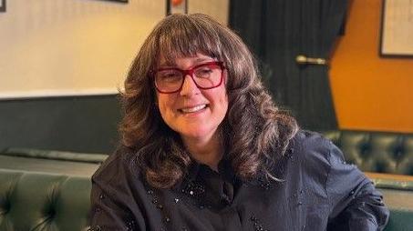Sitting on a green leather banquette, a smiling Jonna Mundy is wearing a pair of bright red glasses and wearing a black blouse