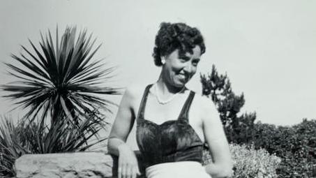 Black-and-white holiday photograph of Mrs Gay looking sideways and grinning while posing against a backdrop of palm trees and shrubbery.