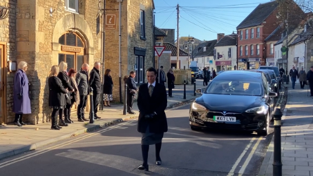 Woman in black bows her head and walks down a small street as a black car follows behind. People stand at the side of the street and clasp their hands. There are buildings either side of the image.