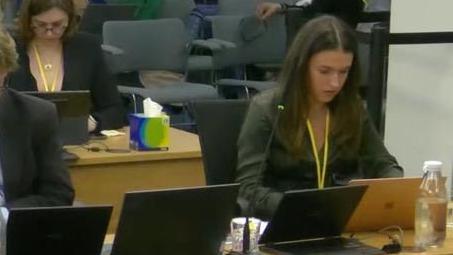 Isabella Thomas sitting at the inquiry with a yellow lanyard round her neck looking at her laptop, with other laptops seen on the desk next to her