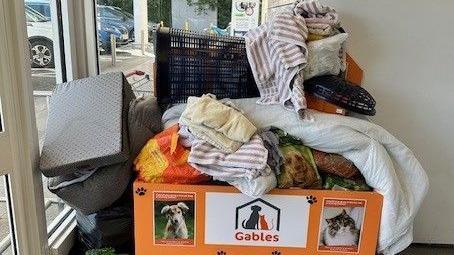 The Gables donation bin which is overfull with items. There is a picture of a dog and a cat on the front and there is a white sign with the Gables log and the word 'Gables' below it.