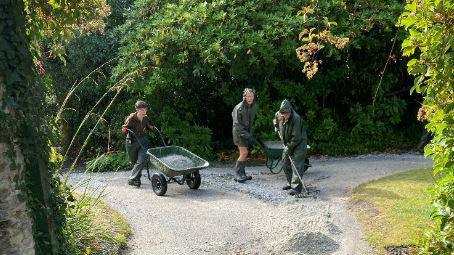 Three people wearing wet weather gear have a wheelbarrow full of grey gravel and are raking gravel onto a path. A full second wheelbarrow is behind them and there are trees and foliage behind and to the side of the image.