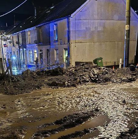 Mud strewn across the Cwmtillery street. The photo is taken from the top of the street, in the foreground there is loads of mud and sludge, and you can see houses towards the back of the picture on a street where there is the flashing blue of the emergency services. 