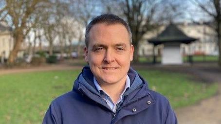 Tom Crone, wearing a blue jacket, stands in a park with a kiosk and trees behind him