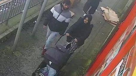 Man in striped puffa jacket stood next to a woman in a dark headscarf pushing a pram on a narrow pavement