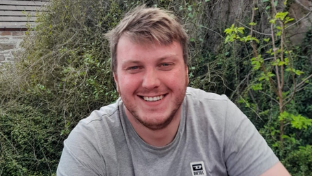 Dylan Boon is sitting outdoors. He has fair hair and stubble on his chin. He is smiling. Dylan is wearing a grey t-shirt.