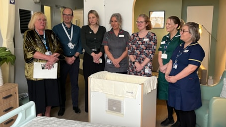 Seven people most in uniform stand with hands clasped in front of them with a cot in front of them.