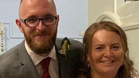 Ed and Christyann Thomas are pictured on their wedding day in October 2021. They are both smiling at the camera. The candles on the church altar are visible in the background. Ed has a white flower in his buttonhole.