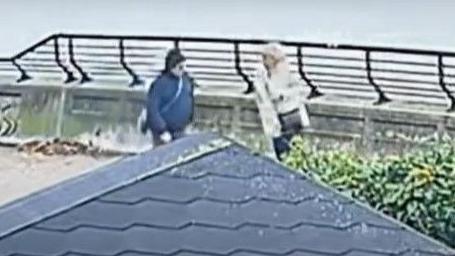 A still image taken from CCTV footage of two people walking along the bank of the Humber estuary. A figure on the right is wearing a dark jacket and what looks to be a black hat. The person on the right of the image has a dark cross body bag, a light coat and light hair.