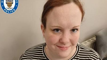 A woman wearing a black and white-striped top sits down and looks up at the camera with a smile. A white wall and radiator are behind her.