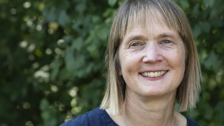 Christine Luxton smiles at the camera while standing in front of a large bush. She has shoulder length blonde hair and a fringe. She wears a navy top.