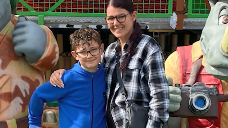 A boy with curly brown hair, glasses and a blue zip-up fleece smiles at the camera next to his mother, who has dyed red hair tied into a long plait and a black and white chequered overshirt. They are standing in front of large plaster models of cartoon characters. 
