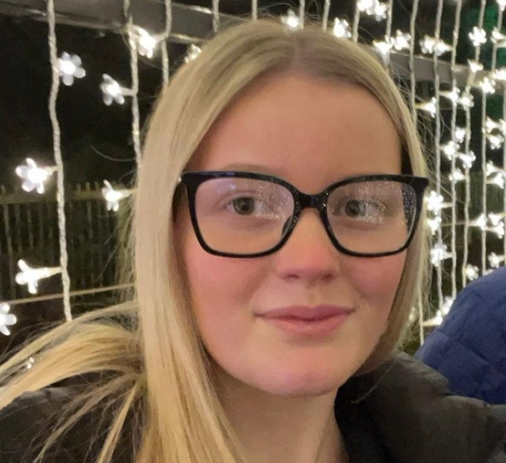 Grace Darnbrough smiles at the camera with fairy lights behind her. She has straight blonde hair and wears black glasses and a black puffer jacket.