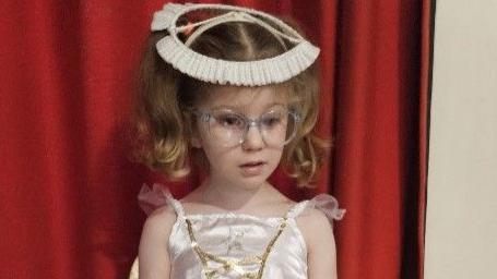 Ella is wearing plastic-framed spectacles. She is wearing her blonde-brown hair in two pigtails with a halo on her head, and a shimmery ivory strappy dress with gold asymmetric lines on the front.