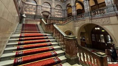 A grand, sweeping marble staircase leads from the front entrance of the town hall upstairs to the council chamber. It has a red carpet running down the middle, with marble bannisters. There are ornate marble arches in the town hall foyer and tall ceilings.