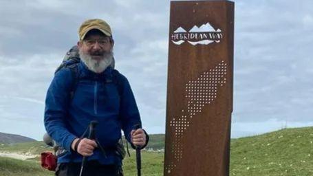 Charles Bain Smith stands beside the Hebridean Way sign in outdor walking gear and holding walking poles, with the Scottish coastline and mountains in the background.