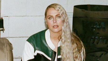 Sally is crouched down beside a generator. She is wearing a green, white and black leather jacket. She has long blonde wavy hair. She is also wearing bright yellow shoes.