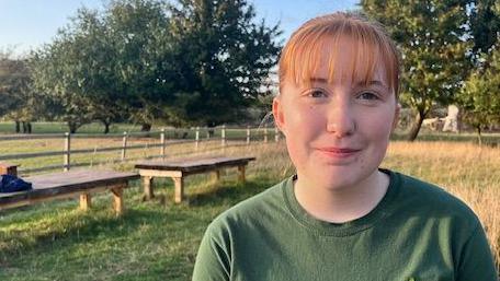 A 14-year-old girl stands in a grassy meadow by two large, wooden benches. In the background is a wooden fence and a row of green trees.