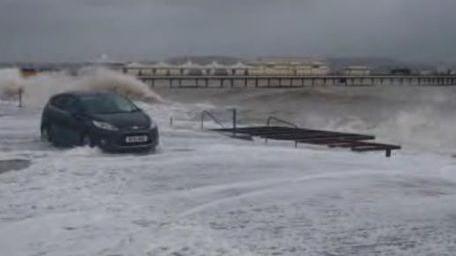 A black car is pounded by the waves on Paignton seafront during Storm Emma in March 2018. You can't see the beach for all the foam. The pier is in the distance set against a stormy grey sky.
