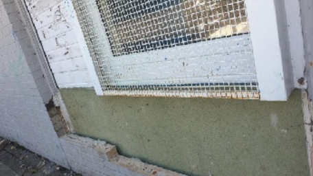 A grey brick wall, with many bricks missing, above a rusty metal window shutter.