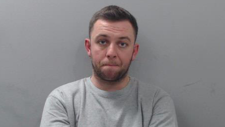 An image of Matthew Brogan sitting in front of a grey wall. He has short brown hair and a beard. He is looking at the camera with a neutral expression on his face. He is wearing a light grey, crewneck jumper.