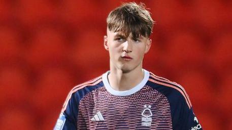 Justin Hanks in action for Nottingham Forest's under-21 side. He is wearing a pink and blue patterned shirt with navy blue short sleeves and three pink stripes running down the shoulders. He is walking and looking into the distance. The background is red showing the empty seats in the stand.