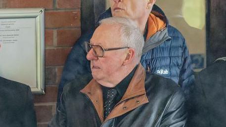 Dennis Taylor at the funeral of snooker player Terry Griffiths. He is wearing a black shirt, a black jacket with brown collar and a black tie with white spots 