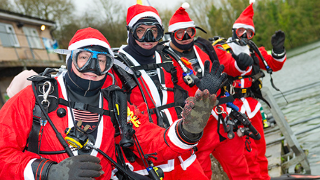 Four scuba divers dressed in Santa outfits waving at the camera at the water's edge