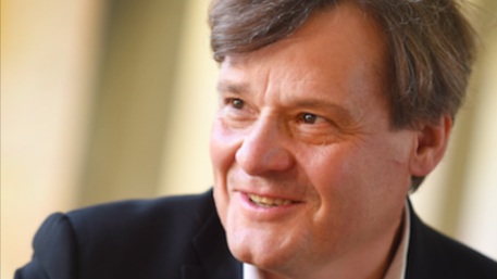 A warmly lit portrait of Dr Wingfield with short, light brown hair, dressed in a dark jacket over a pale shirt. The background is softly blurred.