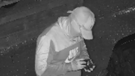 A person in a sports jacket and baseball cap, wearing gloves, peers down at a device. They are outside, with a brick wall behind them. The image is a black and white still from CCTV footage.