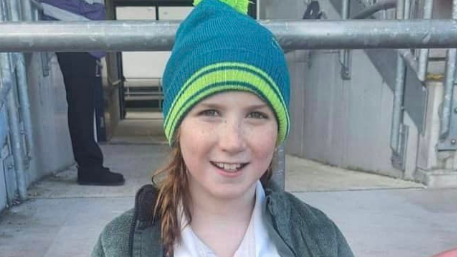 A young girl sits and smiles at the camera holding a white, green and black rugby ball. She is wearing a green and blue beanie hat, a green coat and rugby shirt