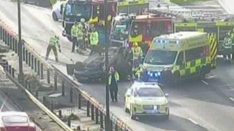 A motorway camera shows a number of emergency services vehicles. Traffic is backing up behind them. A car is on its roof.