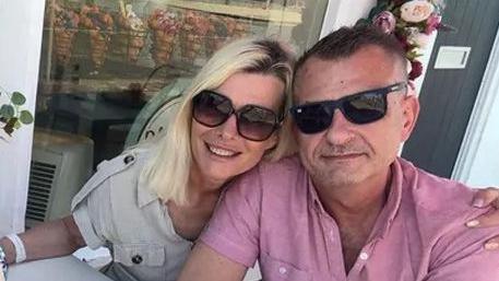 Justyna is hugging her husband Wojtek. They are sitting at a table eating dessert. Wojtek has an ice cream in his hand.  He is wearing a flamingo or watermelon coloured short sleeved shirt. Justyna is wearing an off white top.  Both are wearing sunglasses.