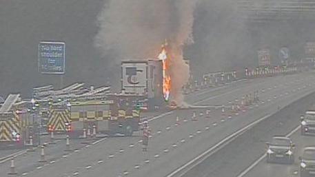 A lorry on fire on the M1 surrounded by fire engines, cones and a fire officer standing in the road. Cars are just visible driving past on the other carriageway