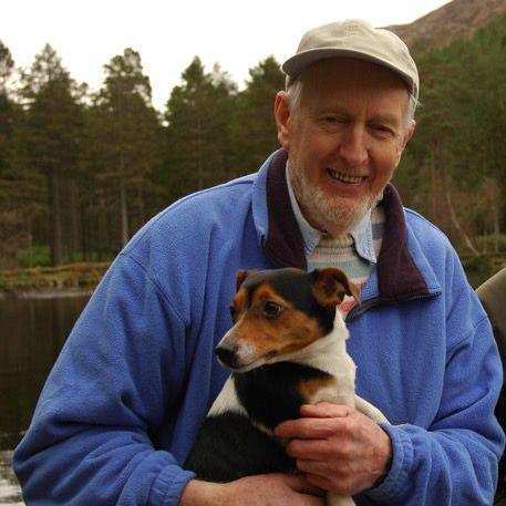 Hamish MacInnes is wearing a cap and a blue fleece over a shirt and striped jumper,. He is smiling and holding a Jack Russell dog. 