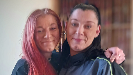 Two women, one with red hair and another with black hair. The woman with red hair has her arm around the shoulder of the other woman. They are both smiling and the background is blurred. They are standing indoors in front of a wooden cupboard.