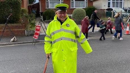 Tony Miles smiles for the camera as he holds his lollipop patrol sign down by his side. He is wearing a long yellow fluorescent raincoat and a matching peak cap.