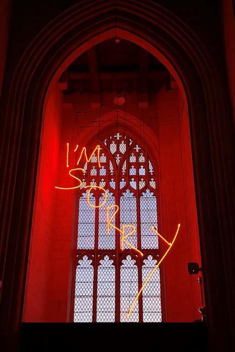 The words "I'm sorry" in neon yellow lettering hanging down with a large church window in the background.