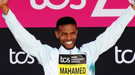 Mahamed Mahamed holding his arms aloft, smiling. He is wearing a white, long-sleeved top. He is standing in front of pink and black banners for the tcs London Marathon. He is wearing a yellow and black sign on his chest saying 'Mahamed'.