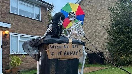 Two skeletons stood by a bin, one is holding an umbrella. There is a sign that says 'Witch bin day is it anyway?'
