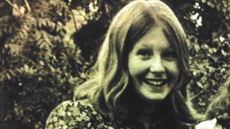 A black and white image of a girl smiling in a garden. She has shoulder-length wavy fair hair and is wearing a floral dress.