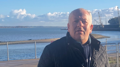 A head and shoulders image of Melven Doe. He is standing outside in front of a harbour and next to some metal fencing. He is wearing a black jacket and a scarf and he is looking directly into the camera.