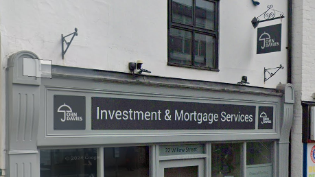 The front of a shop which is painted green grey and has a black and white logo. The logo says "John Davies investment and mortgage services". The wall of the shop, above the sign, is white. There is a black wrought iron hanging sign with a white "John Davies" logo.