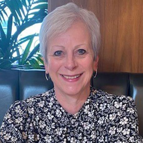 Judith Logan is smiling broadly. She has short grey hair and she is wearing a black, white and grey floral blouse. She has hooped earrings and is sitting on leather seating. Judith Logan understands life-saving surgery must take priority


