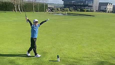 Freddie wearing a light grey cap and blue gold top and black golf pants and white golf shoes throws his arms up in the air to celebrate his hole-in-one on Carus Green Golf Club course.